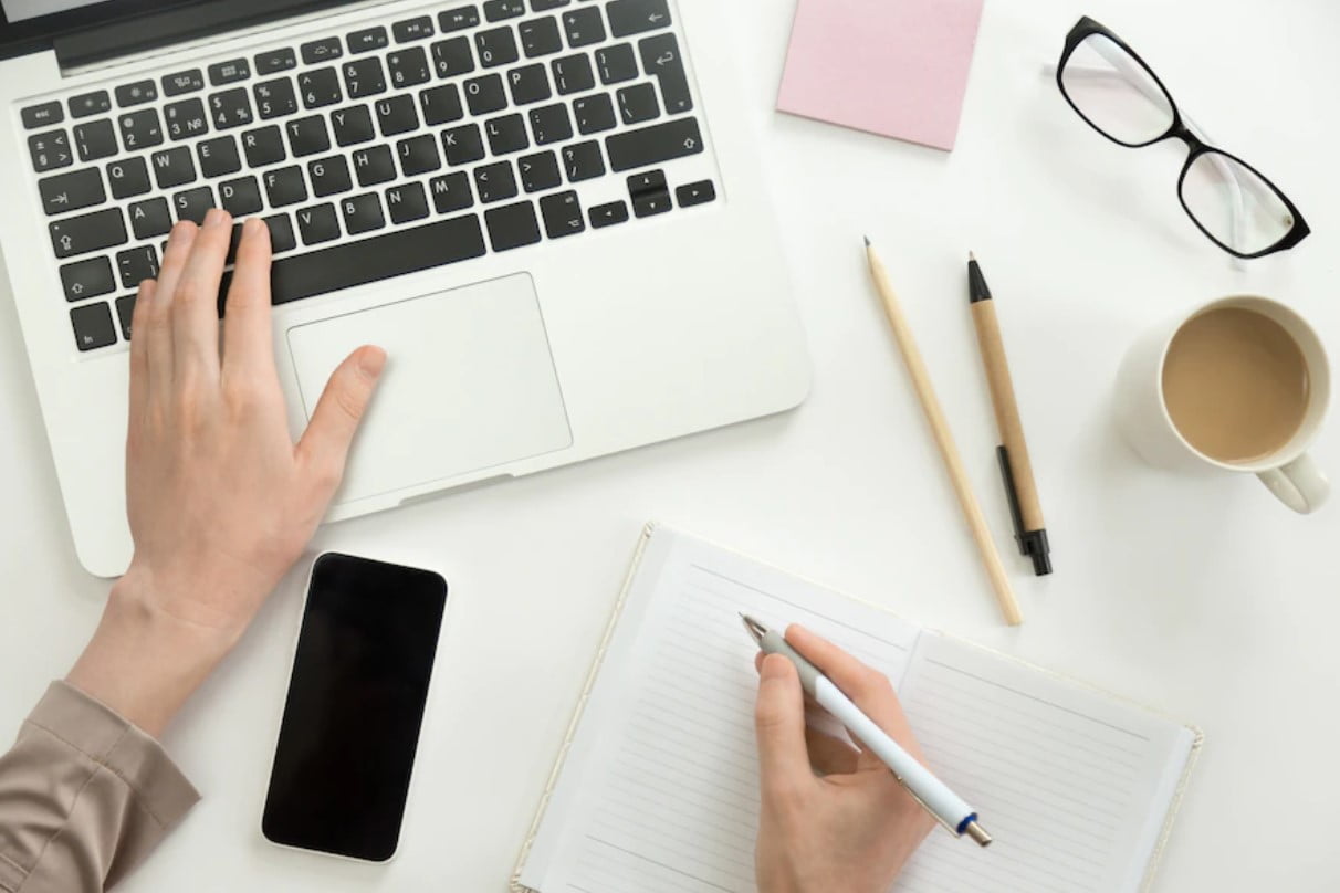 Overhead view of hands typing on a laptop and writing in a notebook, with phone, glasses, coffee cup, and pens on a white desk