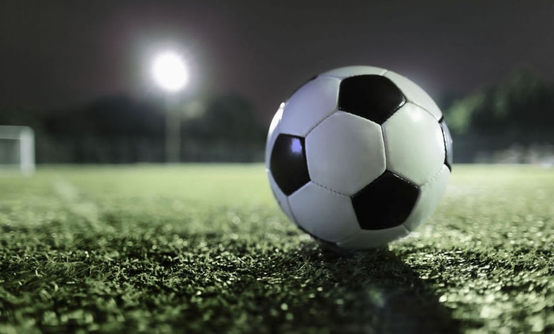 Close-up of a black and white soccer ball on a grass field at night, illuminated by a stadium light